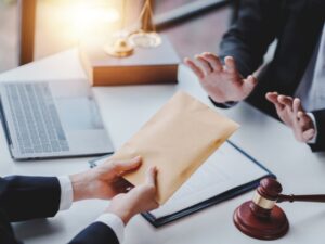 Two suited professionals exchange a manila envelope over a desk with a laptop, legal documents, a gavel, and scales in view, suggesting a legal meeting or filing.