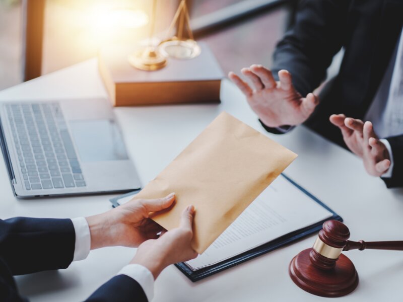 Two suited professionals exchange a manila envelope over a desk with a laptop, legal documents, a gavel, and scales in view, suggesting a legal meeting or filing.