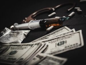 Syringe with a vial and scattered pills beside a stack of US hundred-dollar bills on a dark surface.