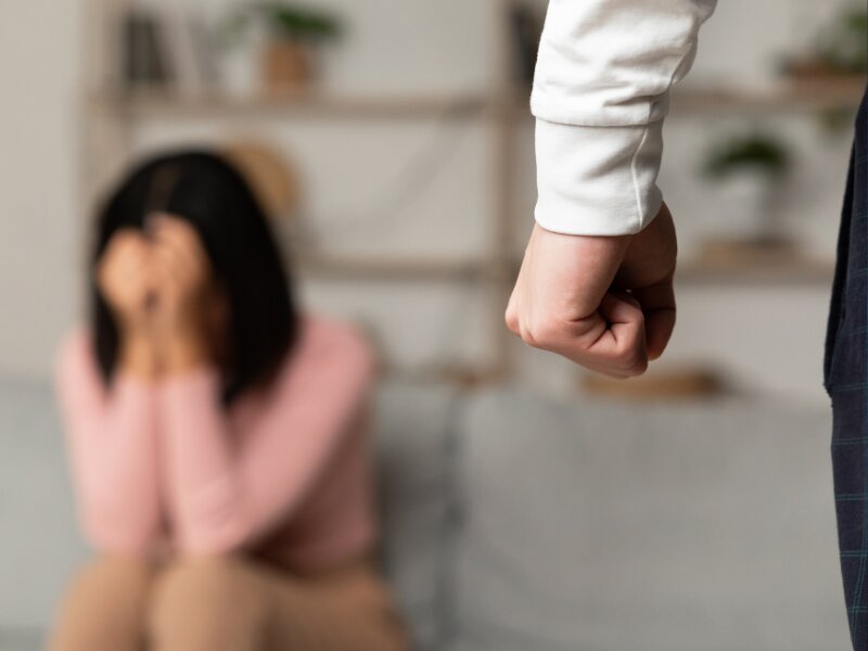 A man’s clenched fist in the foreground toward a distressed woman covering her face in a living room setting.