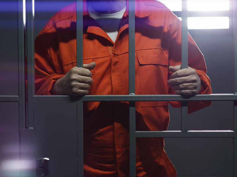 Person in an orange jail jumpsuit grips metal bars in a cell.