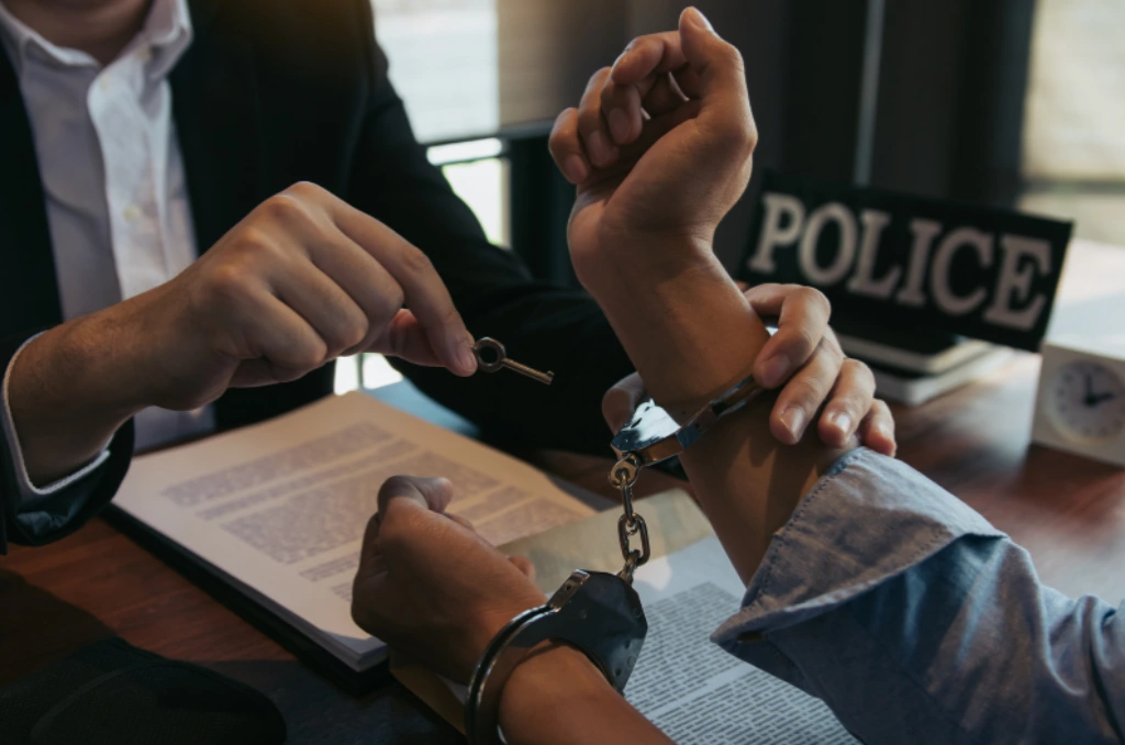 Officer is handcuffing a suspect at a desk; a 'POLICE' sign sits in the background near documents on the table.