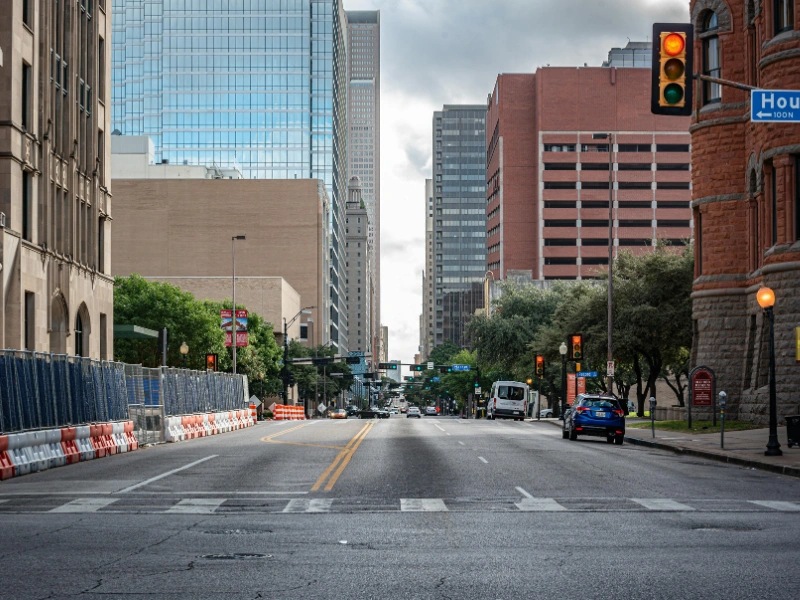 Downtown street scene with construction barriers along the left sidewalk, tall office buildings, and a traffic light at an intersection.