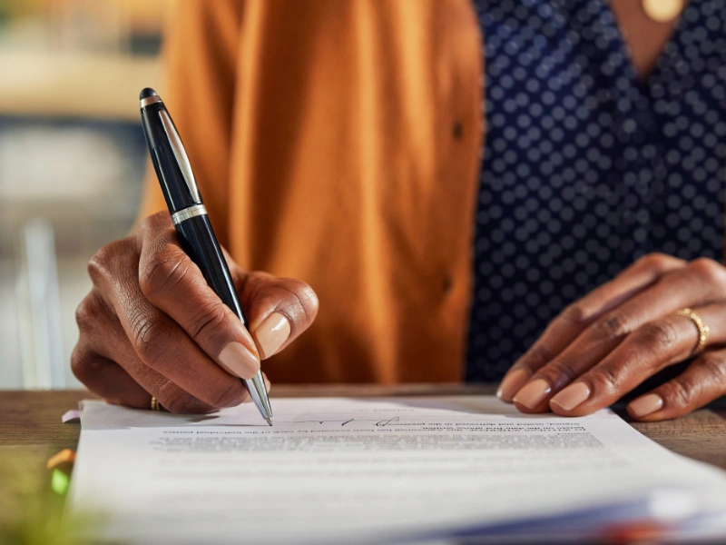 Close-up of a hand signing a document with a black pen on a wooden table.